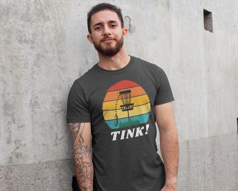 May include: A dark grey t-shirt with a retro-style graphic of a disc golf basket in front of a sunset. The text "TINK!" is printed below the graphic.