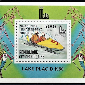 Puede incluir: Un trineo amarillo y negro con dos atletas dentro, corriendo por una pista. El sello es de la República Centroafricana y conmemora los Juegos Olímpicos de Invierno de 1980 en Lake Placid, Nueva York. El sello presenta el texto "Vainqueurs Schaerer-Benz Suisse" y "500f Poste Aerienne".