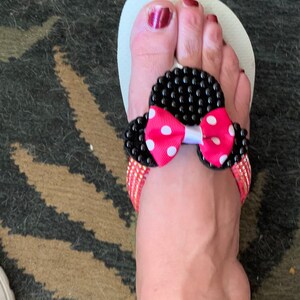 May include: White flip-flop sandals with a black and pink bow embellishment. The bow has white polka dots and black beads around the edges.