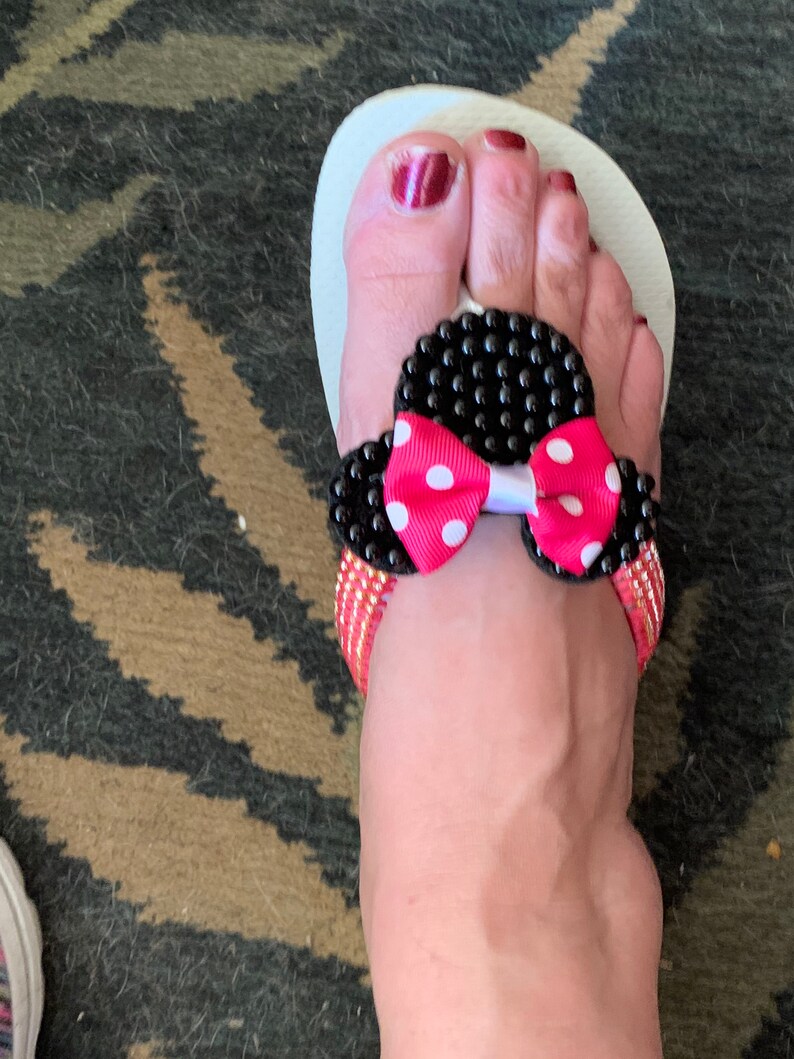 May include: White flip-flop sandals with a black and pink bow embellishment. The bow has white polka dots and black beads around the edges.