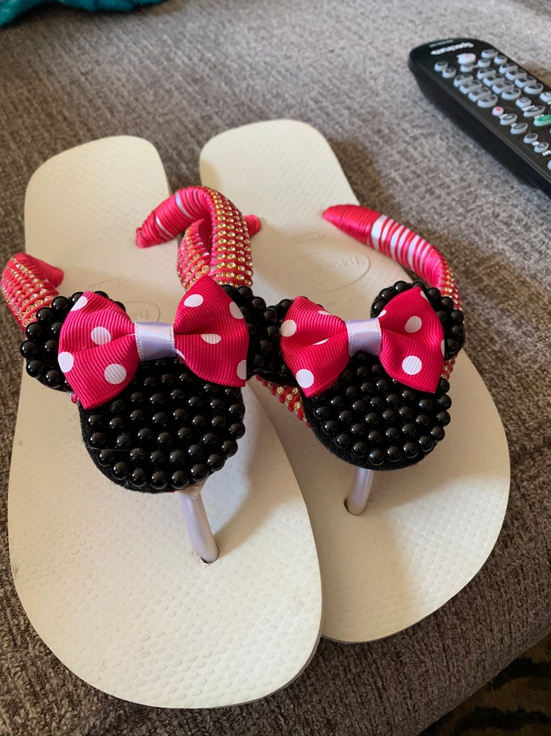 May include: White flip-flops with black and white polka dot bows and black bead embellishments. The bows are shaped like Minnie Mouse ears.