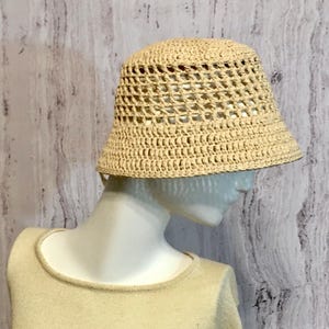 May include: A beige crochet bucket hat with a wide brim. The hat has an open weave design, allowing for ventilation. The hat is displayed on a mannequin head and is a neutral color.