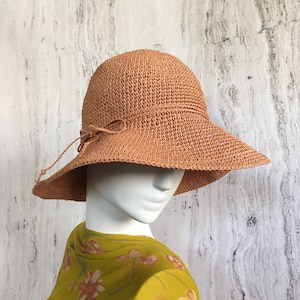 May include: A peach-colored woven sun hat with a wide brim and a decorative bow. The hat is displayed on a mannequin head. The hat has a rounded crown and a chin strap. The hat is made of a woven material.