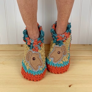 May include: Two crocheted slippers with a cat design. The slippers are multi-colored with orange, teal, yellow, and brown. The cat design is made of brown felt and has a button for an eye.