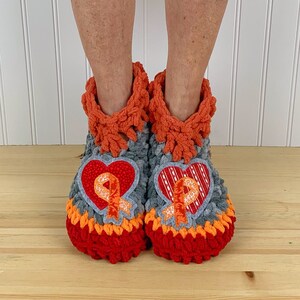 Puede incluir: Un par de zapatillas de crochet grises, naranjas y rojas con un parche en forma de coraz&oacute;n en cada zapatilla. Los parches presentan una cinta naranja.