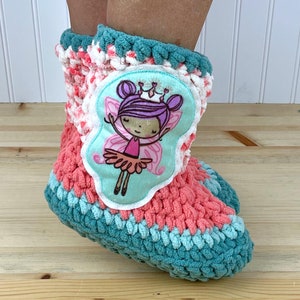 Könnte beinhalten: Türkise, rosafarbene und weiße Häkel-Hausschuhe mit einem Filz-Applikationen einer Fee mit Flügeln, einer Krone und lila Haaren.