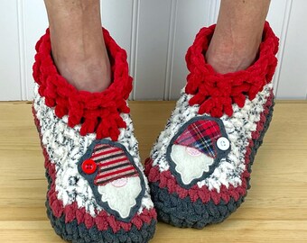 Puede incluir: Un par de zapatillas de crochet grises, blancas y rojas con una aplicación de gnomo en cada una. Las aplicaciones de gnomo tienen un gorro a rayas rojas y blancas, una barba a cuadros y una nariz roja de botón.