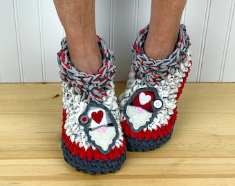 Puede incluir: Un par de zapatillas de crochet grises, rojas y blancas con una aplicación de la cara de Papá Noel. La aplicación tiene un corazón rojo, una barba blanca y una nariz de botón negro.