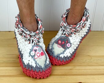 Puede incluir: Un par de zapatillas de crochet en color gris, blanco y rosa. Cada zapatilla tiene una aplicación de fieltro de un gnomo con una nariz de botón rosa y lunares rojos y blancos.