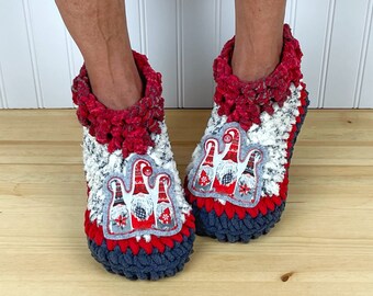 Puede incluir: Pantuflas de crochet rojas, blancas y grises con una aplicación de gnomo. La aplicación de gnomo presenta tres gnomos con gorros rojos y barbas blancas.