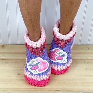 May include: Purple and pink crocheted slippers with a white fuzzy trim. The slippers have a heart-shaped patch with a fairy design and the words "Fairy Magic".