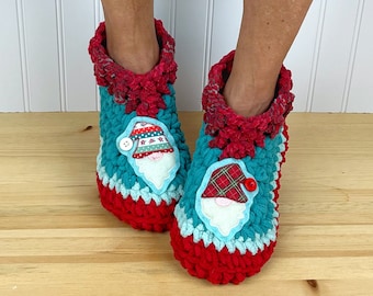 Puede incluir: Pantuflas de crochet rojas y azul verdoso con una aplicación de gnomo. El gnomo tiene un gorro a rayas rojo y blanco, una bufanda a cuadros y una barba blanca.