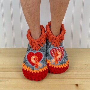 Puede incluir: Un par de zapatillas de crochet grises, naranjas y rojas con un parche en forma de coraz&oacute;n que presenta una cinta naranja. Las zapatillas tienen una textura peluda.
