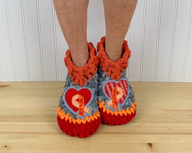 Puede incluir: Un par de zapatillas de crochet grises, naranjas y rojas con un parche en forma de coraz&oacute;n que presenta una cinta naranja. Las zapatillas tienen una textura peluda.