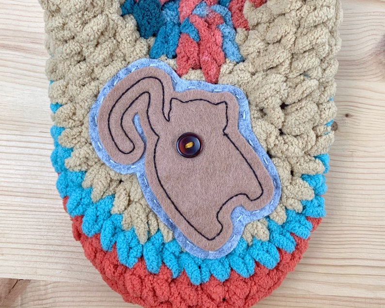 May include: A crocheted bag with a brown felt applique of a cat with a button for an eye. The bag is made with different colored yarn in shades of brown, blue, and orange.