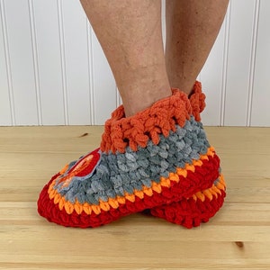 Puede incluir: Un par de zapatillas de crochet en rojo, naranja y gris. Las zapatillas tienen un aspecto texturizado y esponjoso y est&aacute;n hechas con un hilo grueso.