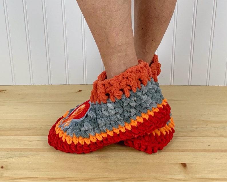 Puede incluir: Un par de zapatillas de crochet en rojo, naranja y gris. Las zapatillas tienen un aspecto texturizado y esponjoso y est&aacute;n hechas con un hilo grueso.