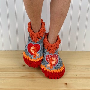 Puede incluir: Un par de zapatillas de crochet con un dise&ntilde;o en forma de coraz&oacute;n que presenta una cinta naranja. Las zapatillas son grises, naranjas y rojas.