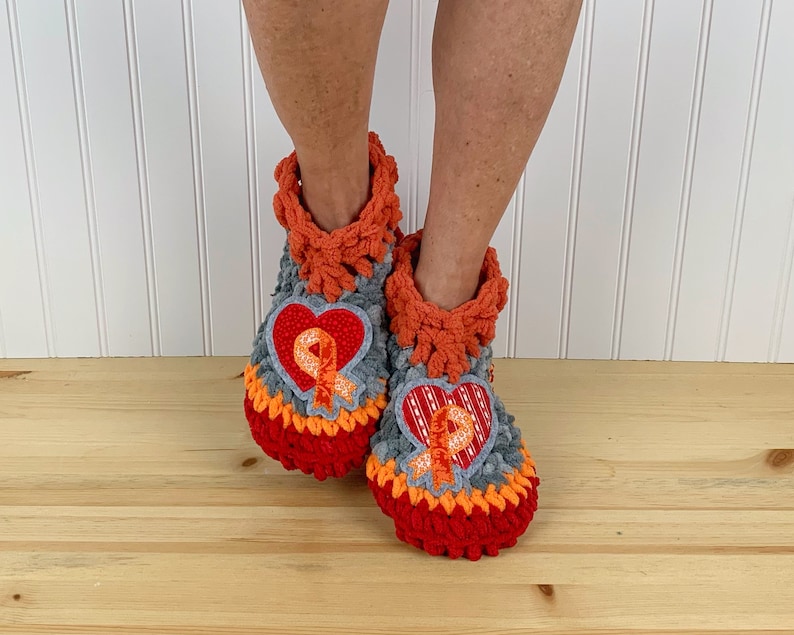Puede incluir: Un par de zapatillas de crochet con un dise&ntilde;o en forma de coraz&oacute;n que presenta una cinta naranja. Las zapatillas son grises, naranjas y rojas.