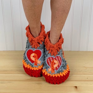 Puede incluir: Un par de zapatillas de crochet en color gris, naranja y rojo. Cada zapatilla tiene una aplicaci&oacute;n en forma de coraz&oacute;n con una cinta naranja.