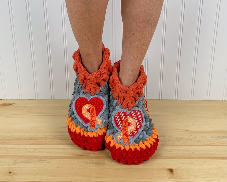 Puede incluir: Un par de zapatillas de crochet en color gris, naranja y rojo. Cada zapatilla tiene una aplicaci&oacute;n en forma de coraz&oacute;n con una cinta naranja.