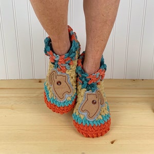 May include: Two crocheted slippers with a brown felt applique of a dog's face on the front. The slippers are multi-colored with orange, teal, and yellow yarn. The slippers are on a wooden floor.