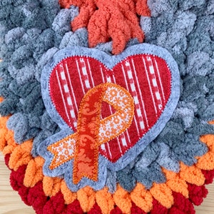 Puede incluir: Un adorno de coraz&oacute;n de ganchillo gris, naranja y rojo con un coraz&oacute;n a rayas rojas y blancas y una cinta naranja con un patr&oacute;n floral.