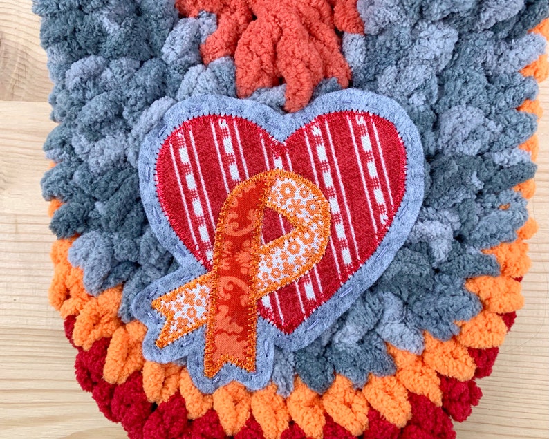 Puede incluir: Un adorno de coraz&oacute;n de ganchillo gris, naranja y rojo con un coraz&oacute;n a rayas rojas y blancas y una cinta naranja con un patr&oacute;n floral.