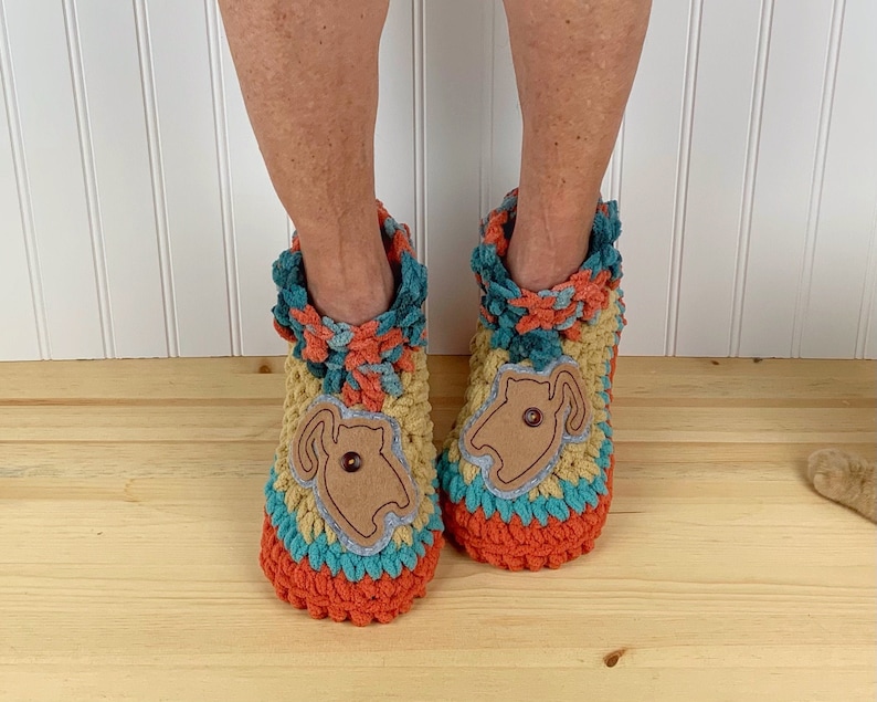 May include: Two crocheted slippers with a cat design. The slippers are multi-colored with orange, teal, yellow, and brown. The cat design is made of brown felt and has a button for an eye.