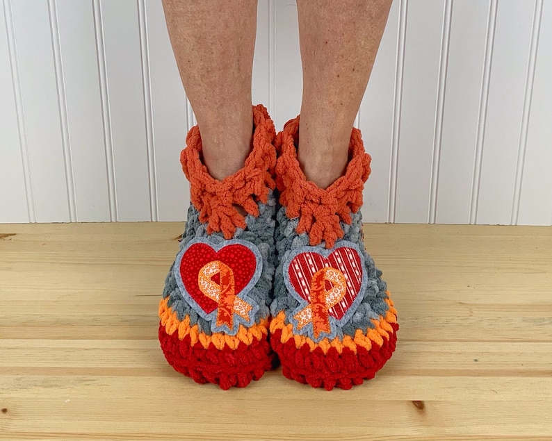 Puede incluir: Un par de zapatillas de crochet grises, naranjas y rojas con un parche en forma de coraz&oacute;n en cada zapatilla. Los parches presentan una cinta naranja.