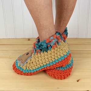 May include: A pair of crocheted slippers with a brown bear applique. The slippers are multi-colored with orange, teal, and tan yarn. The slippers have a crocheted cuff and are made with a soft, fuzzy yarn.