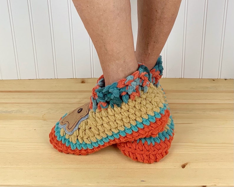 May include: A pair of crocheted slippers with a brown bear applique. The slippers are multi-colored with orange, teal, and tan yarn. The slippers have a crocheted cuff and are made with a soft, fuzzy yarn.