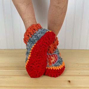 Puede incluir: Pantuflas de crochet rojas, naranjas y grises con un dise&ntilde;o en forma de coraz&oacute;n. Las pantuflas se llevan en los pies de una persona.