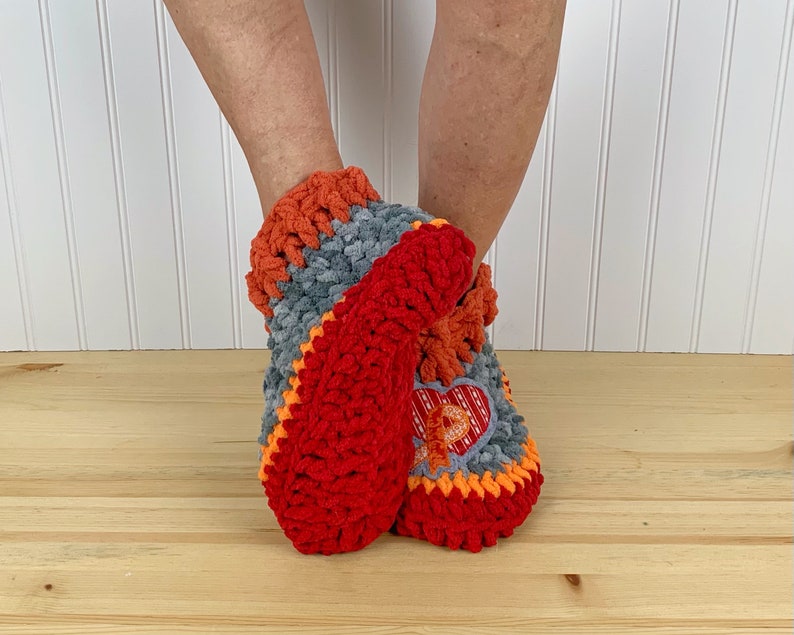 Puede incluir: Pantuflas de crochet rojas, naranjas y grises con un dise&ntilde;o en forma de coraz&oacute;n. Las pantuflas se llevan en los pies de una persona.