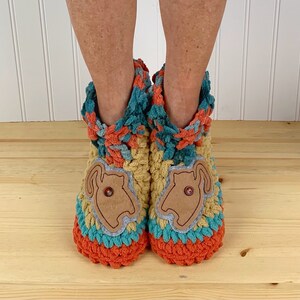 May include: A pair of crocheted slippers with a cat design on the front. The slippers are made in a multi-colored yarn with shades of orange, yellow, blue, and green. The cat design is made from a brown fabric and has a button for an eye.