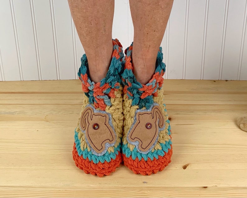 May include: A pair of crocheted slippers with a cat design on the front. The slippers are made in a multi-colored yarn with shades of orange, yellow, blue, and green. The cat design is made from a brown fabric and has a button for an eye.
