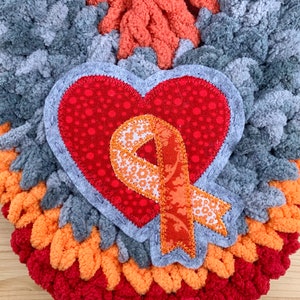 Puede incluir: Un tapiz de pared de forma de coraz&oacute;n tejido a crochet en gris, naranja y rojo con una aplicaci&oacute;n de coraz&oacute;n rojo y una cinta naranja.