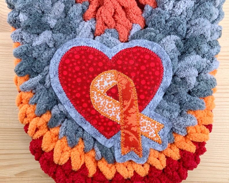 Puede incluir: Un tapiz de pared de forma de coraz&oacute;n tejido a crochet en gris, naranja y rojo con una aplicaci&oacute;n de coraz&oacute;n rojo y una cinta naranja.