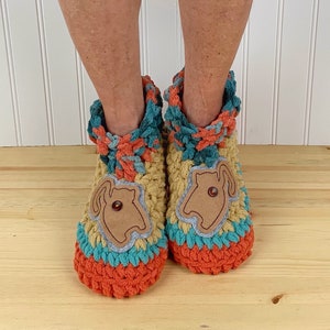 May include: Two crocheted slippers with a brown felt applique of a cat on each. The slippers are orange, teal, and yellow. The slippers are on a wooden surface.