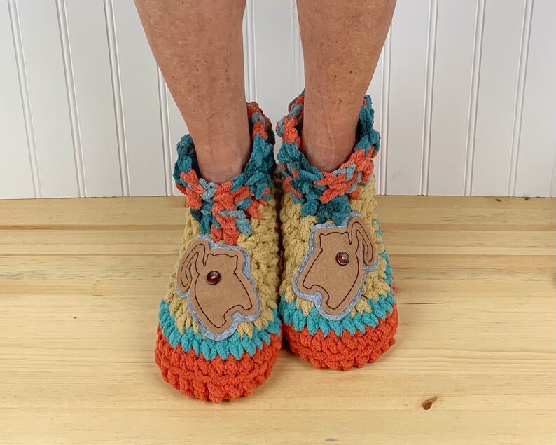 May include: Two crocheted slippers with a brown felt applique of a cat on each. The slippers are orange, teal, and yellow. The slippers are on a wooden surface.