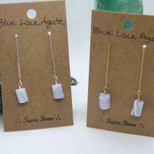 May include: Two pairs of blue lace agate dangle earrings. The earrings on the left have silver chains and the earrings on the right have gold chains. Both pairs of earrings have the text "Sapira Stones" on the card.