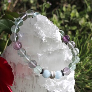 May include: A bracelet made with green, purple, and blue gemstone beads. The bracelet is on a white crystal.