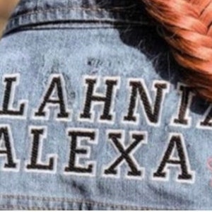 May include: A blue denim jacket featuring the embroidered text MALAHNIA ALEXA in black letters with white outlines. To the left of the text is a small rainbow-colored circular emblem, and to the right is a small pink five-petaled flower outline.