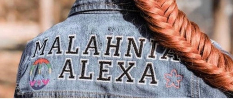 May include: A blue denim jacket featuring the embroidered text MALAHNIA ALEXA in black letters with white outlines. To the left of the text is a small rainbow-colored circular emblem, and to the right is a small pink five-petaled flower outline.