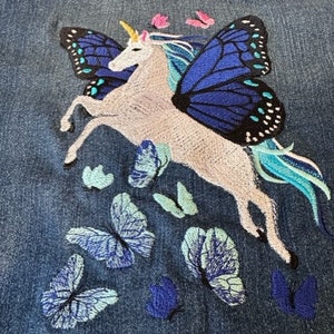 May include: A blue denim jacket featuring an embroidered design of a white unicorn with large blue and black butterfly wings. The unicorn has a yellow horn and a flowing teal mane and tail, surrounded by several smaller embroidered butterflies.
