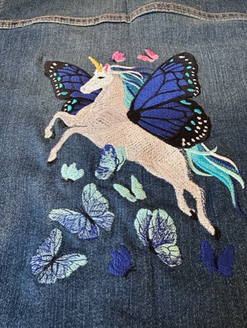 May include: A blue denim jacket featuring an embroidered design of a white unicorn with large blue and black butterfly wings. The unicorn has a yellow horn and a flowing teal mane and tail, surrounded by several smaller embroidered butterflies.