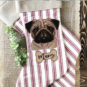 May include: A personalized Christmas stocking with a red and white striped pattern. The stocking features a brown bone-shaped tag with the name "Mavis" written on it. A pug dog's face is embroidered on the stocking.