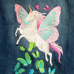 May include: A blue denim fabric featuring an embroidered white unicorn with large pink and teal butterfly wings. The unicorn has a golden horn and a pink mane. Several small green, teal, and pink butterflies are embroidered below and above.