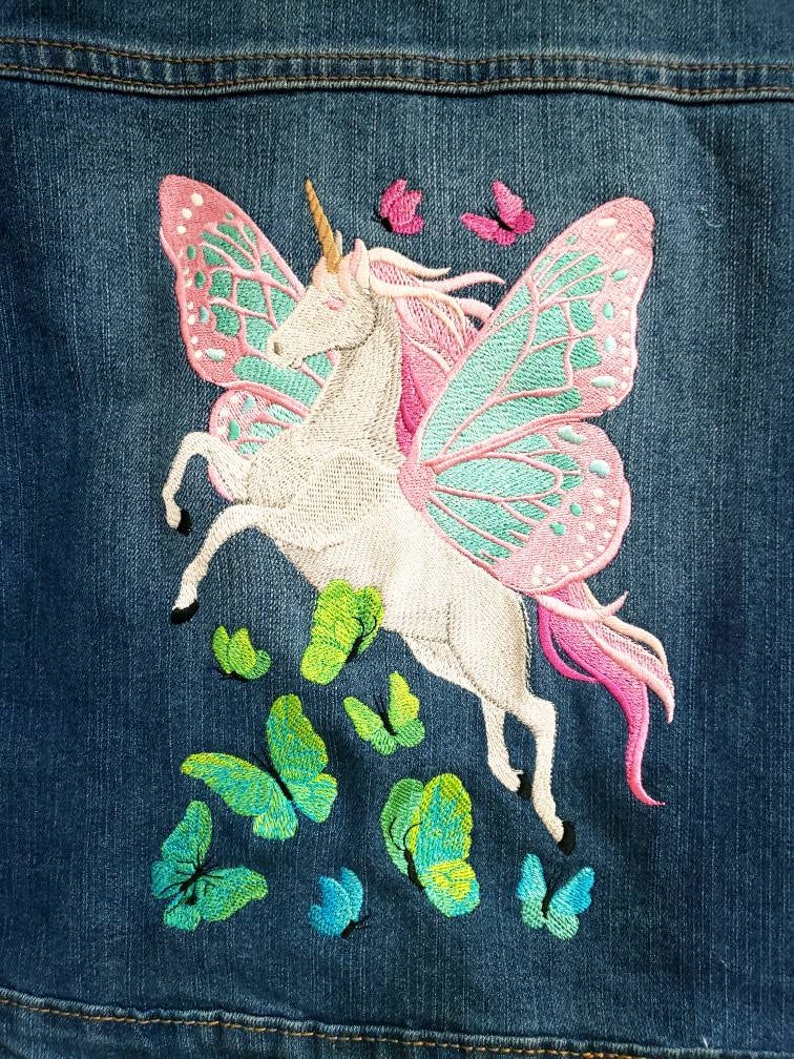 May include: A blue denim fabric featuring an embroidered white unicorn with large pink and teal butterfly wings. The unicorn has a golden horn and a pink mane. Several small green, teal, and pink butterflies are embroidered below and above.