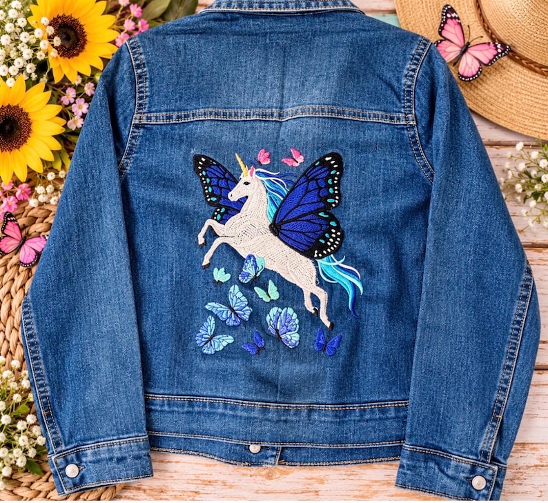 May include: A blue denim jacket featuring a large embroidered design on the back. The embroidery depicts a white unicorn with blue butterfly wings, surrounded by several smaller blue and pink butterflies. The jacket has long sleeves and buttons.